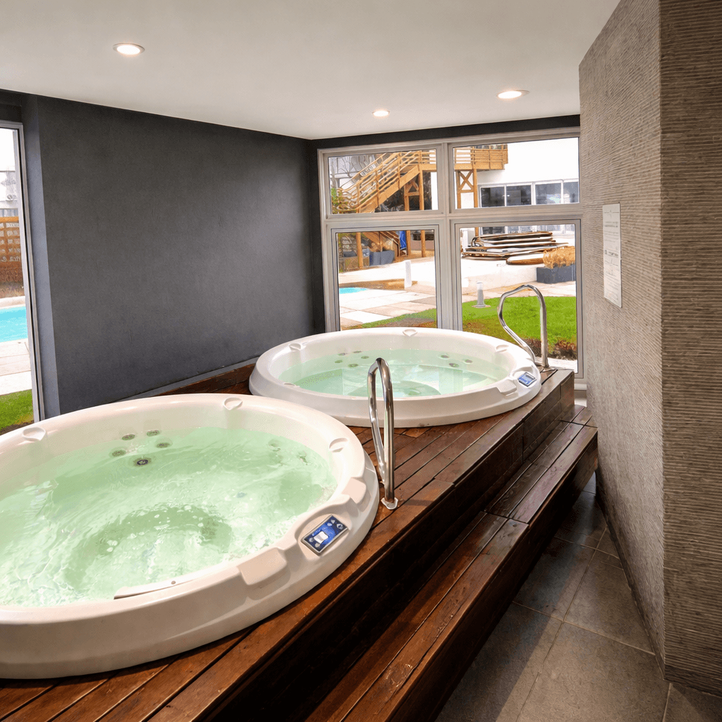 Área de spa con dos jacuzzis interiores, grandes ventanales y vista hacia la piscina exterior. El espacio cuenta con deck de madera, iluminación interior y zona de descanso.