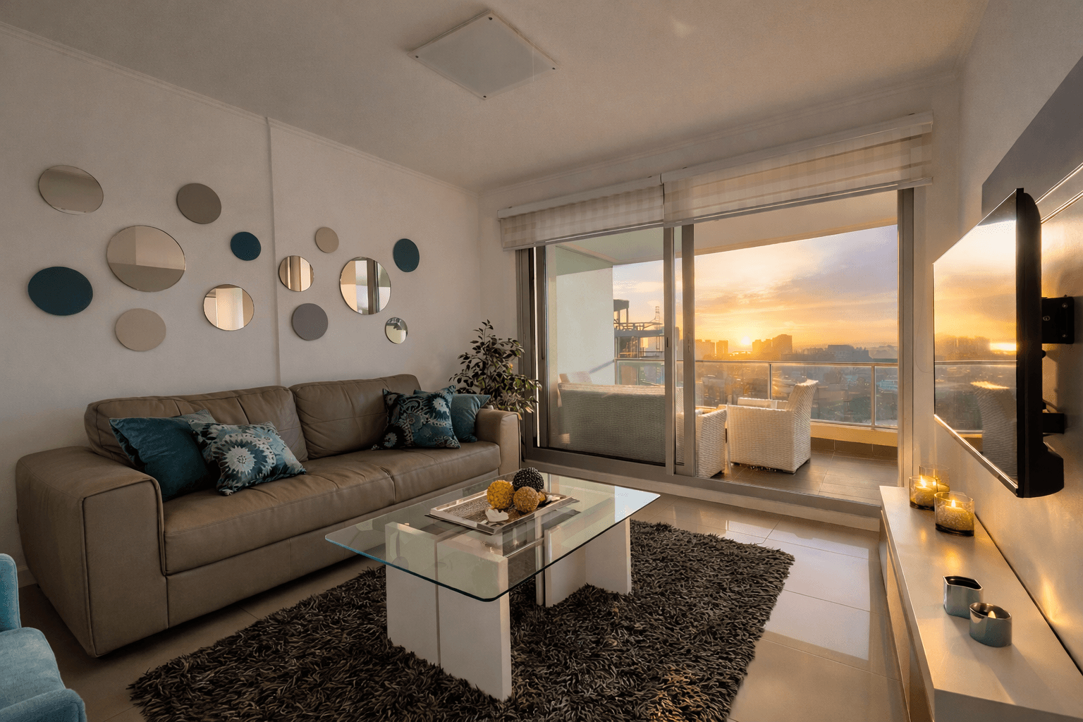 Living moderno y luminoso con sofá, mesa ratona de vidrio y TV, conectado a un amplio balcón con sillones y vista panorámica a la ciudad. Grandes ventanales permiten disfrutar de abundante luz natural y del atardecer.