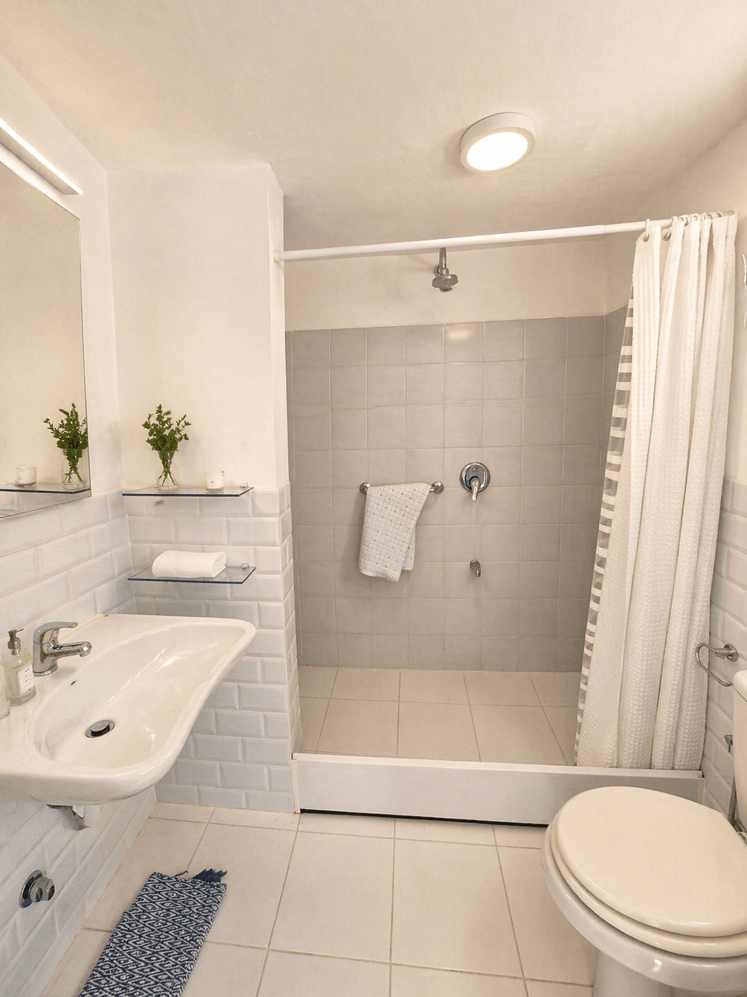 Baño completo y luminoso con ducha, lavabo, inodoro y espejo. La zona de ducha cuenta con cortina y revestimiento de azulejos en tonos claros.