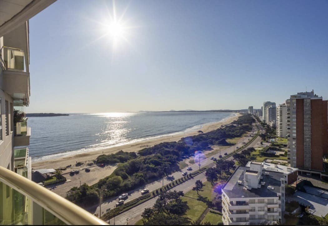 Vista panorámica al mar desde un balcón en Punta del Este, con amplia playa de arena, rambla costera y edificios frente al mar. La imagen muestra una vista despejada de la costa y el océano.