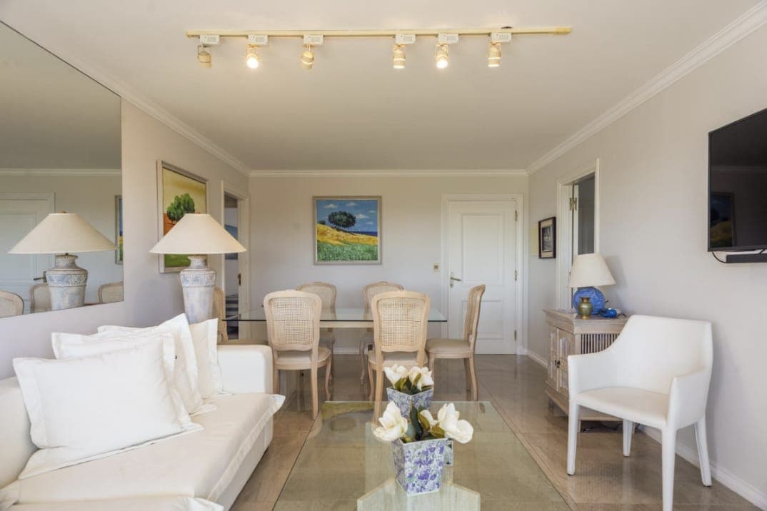 Living comedor amplio y luminoso con sofá, sillones y mesa de comedor. Cuenta con TV, aire acondicionado, grandes espejos y una decoración en tonos claros.
