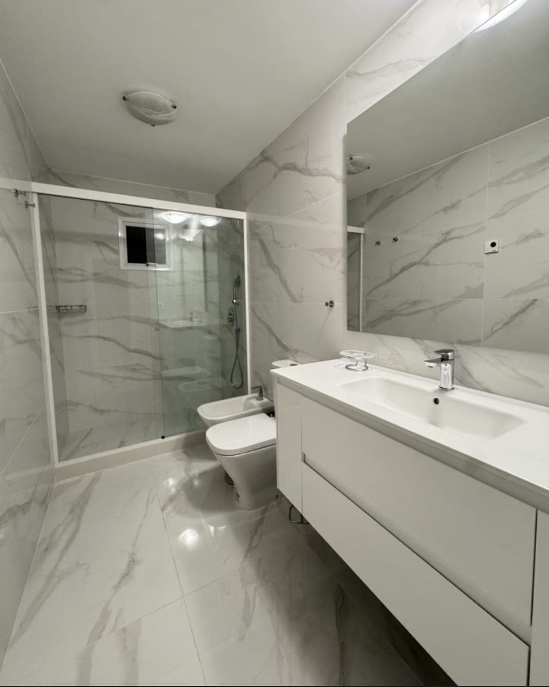 Baño completo con revestimiento efecto mármol, ducha con mampara de vidrio, lavabo con amplia mesada, espejo, inodoro y bidé. Cuenta con mueble bajo mesada para espacio de guardado.
