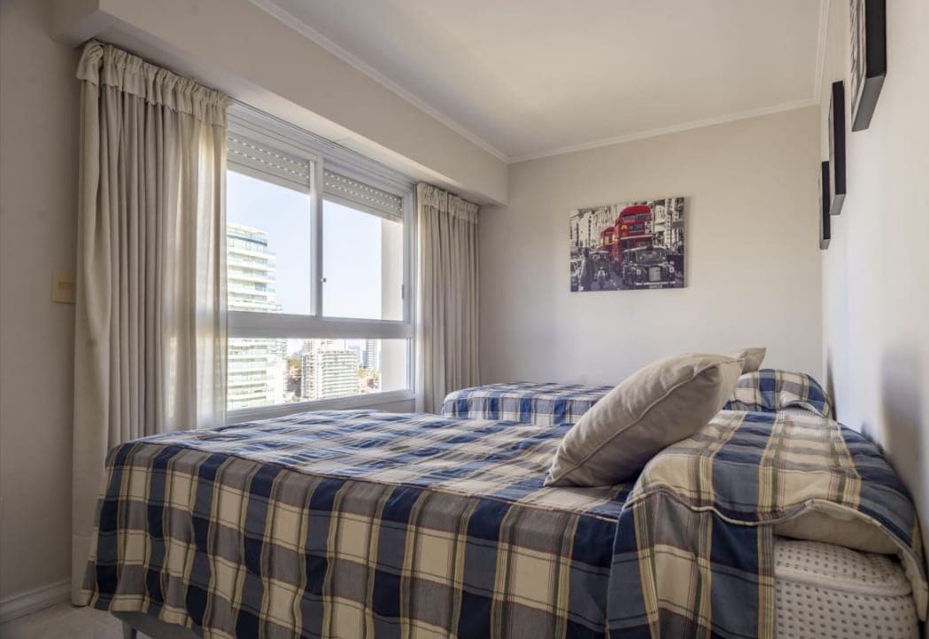 Dormitorio luminoso con dos camas individuales, amplio ventanal y vista urbana. Cuenta con cortinas y decoración en tonos claros.