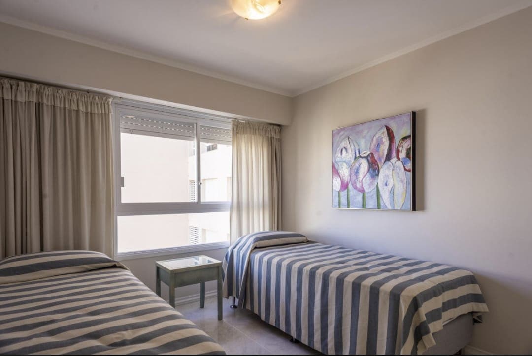 Dormitorio luminoso con dos camas individuales, amplio ventanal y cortinas. Cuenta con mesa de luz entre ambas camas y decoración en tonos claros.
