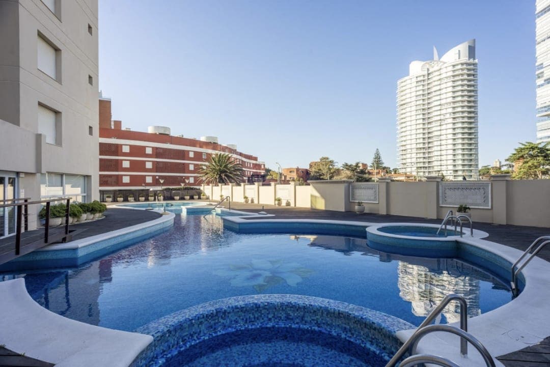 Piscina exterior del edificio con amplias áreas de agua y sectores diferenciados, rodeada de solárium y edificios residenciales. Espacio al aire libre con buena exposición al sol.