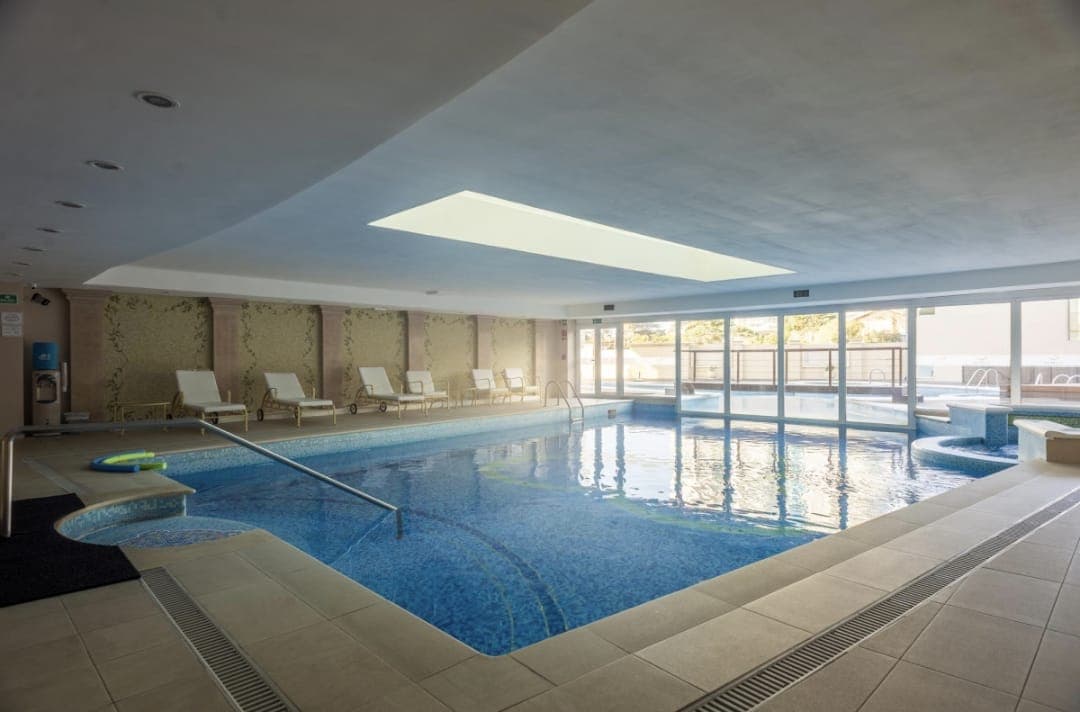 Piscina interior climatizada de edificio con amplio espacio de descanso, reposeras y grandes ventanales que aportan luz natural. Cuenta con diferentes sectores de piscina y acceso mediante escaleras con pasamanos.